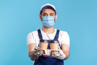 Teslimat servisi. Mavi tulumlu, maske takan, güvenlik eldiveni takan tek kullanımlık bardaklarda içki veren ve kameraya gülümseyerek bakan dost canlısı mutlu kurye. Kapalı çekim, izole edilmiş.