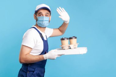Teslimat servisi. Tulumlu, maske takan, elinde kahve ve pizza kutusu tutan, güvenlik eldiveni takan içecek sunan ve kameraya el sallayan sevimli bir kurye. Mavi zemin üzerinde kapalı çekim izole