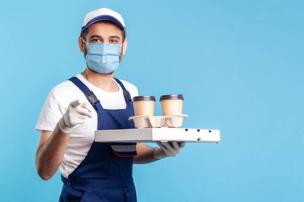 Delivery service. Smiling happy courier in overalls and mask holding coffee, pizza box, wearing safety gloves and offering drinks food, pointing finger to camera, choosing you. indoor shot, isolated