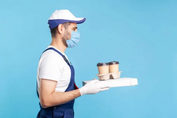Delivery service. Side view, friendly professional courier in uniform and mask holding coffee and pizza box, wearing safety gloves offering drinks and fast food, express shipping. indoor shot isolated