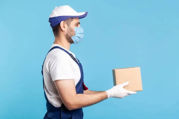 Delivery and cargo transportation. Side view, professional loader man in workwear and mask holding cardboard box. Courier carrying parcel. Online express shipping, relocation moving services. indoor