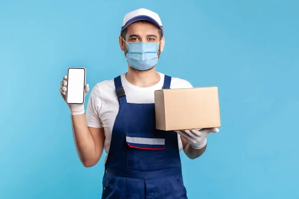 Cargo tracking mobile app. Courier man in uniform and mask holding cardboard box and cellphone with blank display, mock up for express shipping advertise, online delivery order. indoor shot isolated