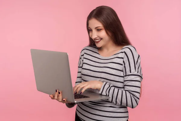 フリーランスの仕事インターネット通信 キーボードのラップトップと笑顔に入力ストライプのスウェットシャツで幸せな若い女性の肖像画 オンラインでチャット ピンクの背景に隔離された屋内スタジオで — ストック写真