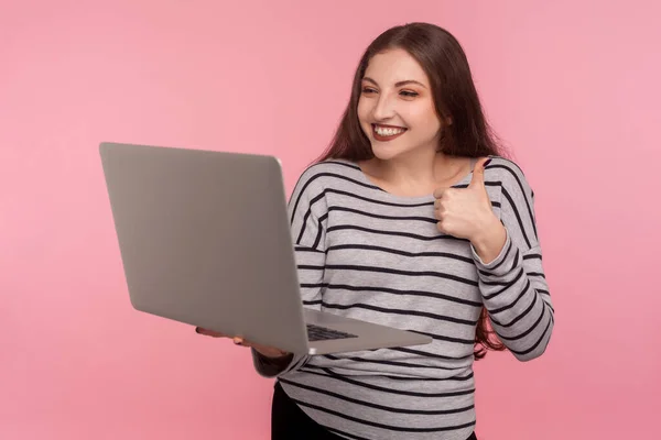 Good job! Portrait of happy woman in striped sweatshirt showing thumbs up to laptop screen, gesturing like, communicating via video call, online conference. studio shot isolated on pink background