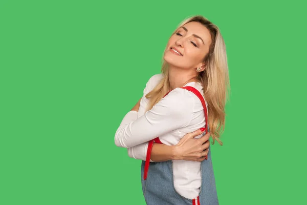 I am beautiful, love myself! Portrait of happy adult blond woman in denim overalls embracing herself and smiling with pleased satisfied expression. indoor studio shot isolated on green background