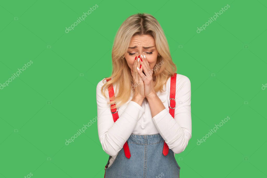 Portrait of upset adult woman in denim overalls crying from sadness, suffering depression and loneliness, negative emotions, frustrated by misfortune. indoor studio shot isolated on green background