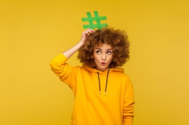 Popüler blog yazıları, moda içeriği. Şaşkın kıvırcık saçlı, şehir tarzı kapüşonlu bir kadının portresi kafasında hashtag ile şaşkınlıkla bakıyor. Stüdyo çekimi sarı arkaplanda izole edildi