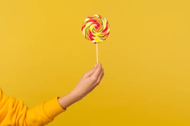 Closeup of hand holding appetizing round rainbow candy on stick, big lollipop in arm, confectionery advertising, glucose sweet foods, sugary dessert. indoor studio shot isolated on yellow background clipart