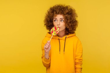 Dalgın kıvırcık saçlı kadın portresi şehir tarzı kapüşonlu, lolipop yalayan, tatlı gökkuşağı şekerlerinin tadını çıkaran ve düşünceli bir ifadeyle başka yere bakan. Stüdyo çekimi sarı arkaplanda izole edildi