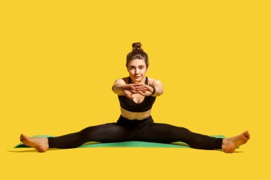 Sıkı spor kıyafetleri içinde, saçları topuz gibi olan formda bir jimnastikçi kadın açık bacaklı ve ellerini uzatarak oturuyor, ısınıyor, spor esnekliği egzersizleri yapıyor. Stüdyo çekimi sarı arkaplanda izole edildi