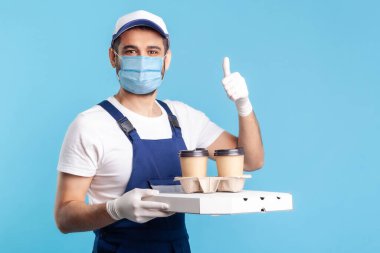 Teslimat servisi. Tulumlu, maske takan, kahve ve pizza kutusu tutan, güvenlik eldiveni takan, yiyecek sunan ve el hareketi yapan dost canlısı iyimser kurye. Kapalı, izole.