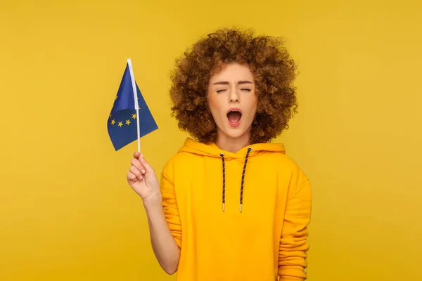 Civil voting rights, EU law. Portrait of curly-haired young woman in hoodie holding European Union flag and keeping her mouth wide open, loudly declaring her rights. indoor studio shot, isolated