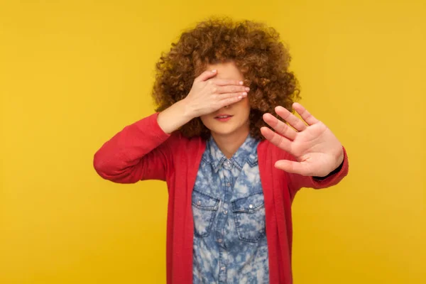 No, don\'t want to look. Portrait of scared woman with curly hair covering eyes and showing stop gesture, feeling shy afraid to watch, ignoring troubles. studio shot isolated on yellow background