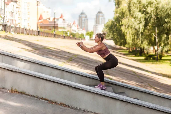 Personal training program. Sporty athletic woman in tracksuit squatting doing sit-ups on stairs outdoor, warming up before jogging, exercising for weight loss, fat burning. Cityscape on background