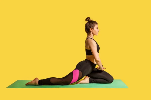 动机严肃的女运动员在垫子上练习瑜伽 做一条腿的王鸽姿势 热身伸展肌肉 做柔韧运动 演播室拍摄 在黄色背景下隔离的运动训练 — 图库照片