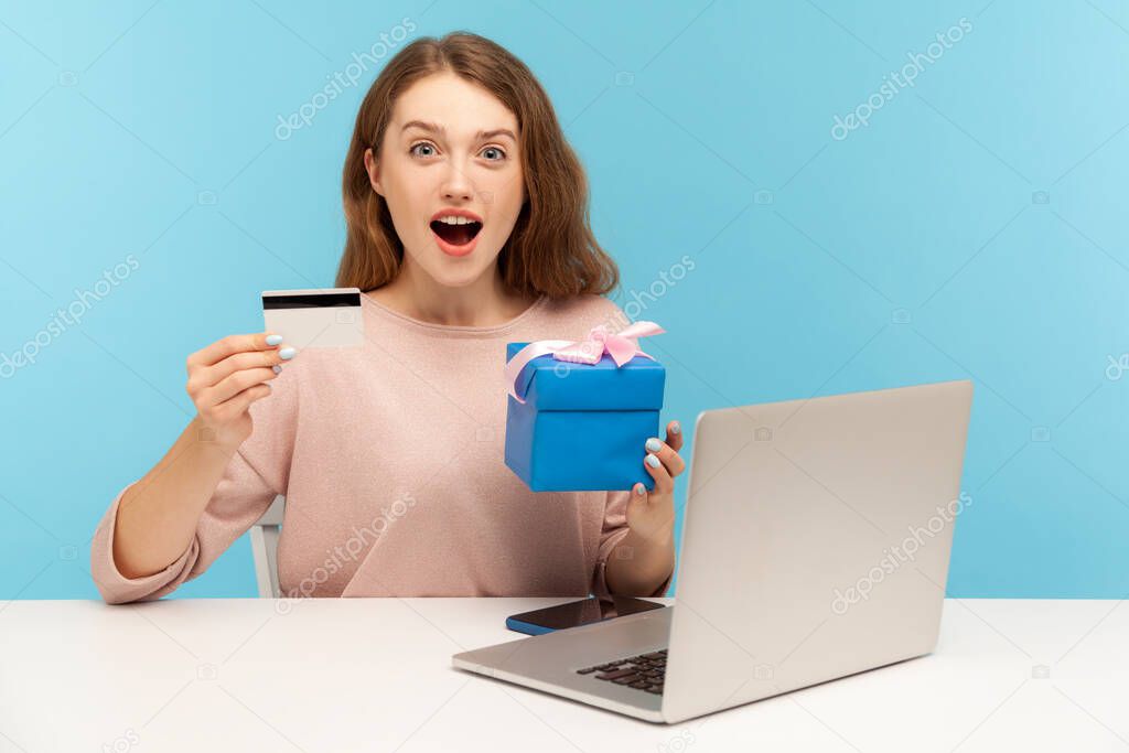 Surprised excited beautiful woman sitting at desk with laptop and holding gift box, showing credit card and looking at camera with amazement, shocked expression. indoor studio shot, blue background