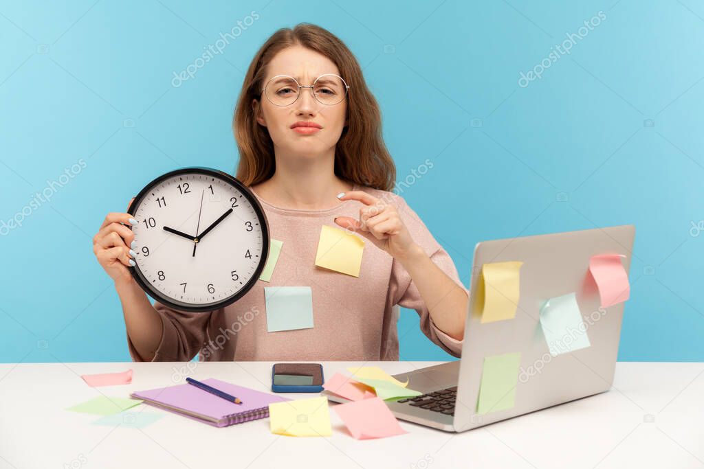 I need some break! Exhausted fatigued upset woman employee sitting at workplace office, all covered with sticky notes, holding big clock and showing a little bit gesture, begging more time. indoor
