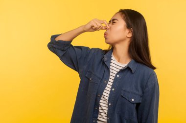 Berbat kokuyor! Tişörtü iğrenç kokan, nefesini tutan, burnu parmaklarıyla tutan, pis kokudan ve osuruktan kaçan bir kızın portresi. Sarı arka planda izole edilmiş kapalı stüdyo çekimi