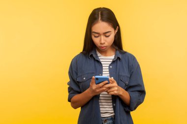 Portrait of attentive serious girl in stylish denim shirt typing message, dialing number calling on cell phone, using online mobile application, searching web. indoor studio shot, yellow background clipart