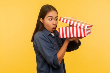 Portrait of amazed funny girl in denim shirt opening gift and looking into box with shocked astonished expression, found unbelievable present surprise. indoor studio shot isolated on yellow background clipart