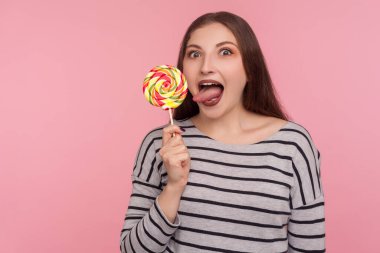 Dilini uzatan komik bir kadının portresi lolipop yalıyor, tatlı, yuvarlak gökkuşağı şekeri tadıyor, lezzetli tatlıların tadını çıkarıyor, tatlı şekerlemeler yapıyor. Pembe arka planda kapalı stüdyo çekimleri