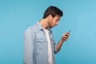 Yan görünüş, işçi kot gömleği giyen, cep telefonundan şok edici bir mesaj okuyan, beklenmedik bir haberle şaşıran, online sohbet için mobil uygulamayı kullanan bir adam. Mavi arka planda kapalı stüdyo çekimleri