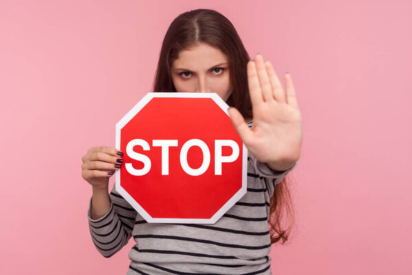 No, prohibited! Portrait of strict bossy girl looking angrily and showing stop gesture, holding road traffic sign as warning of ban, forbidden access. indoor studio shot isolated on pink background