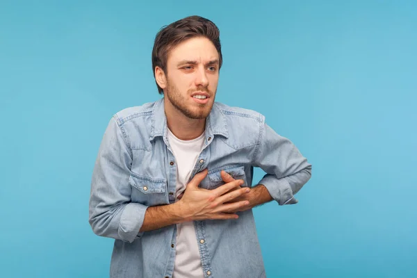 Cardiac problems. Portrait of stressed out, worried man in denim shirt clutching chest, suffering heart attack, needs emergency of acute myocardial infarction. studio shot isolated on blue background
