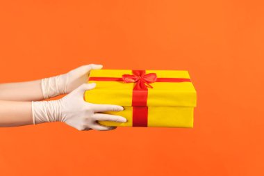 Profile side view closeup of human hand in white surgical gloves holding yellow gift box. indoor, studio shot, isolated on orange background.