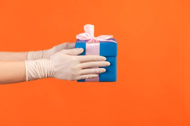 Profile side view closeup of human hand in white surgical gloves holding blue gift box. sharing, giving or delivery concept. indoor, studio shot, isolated on orange background.