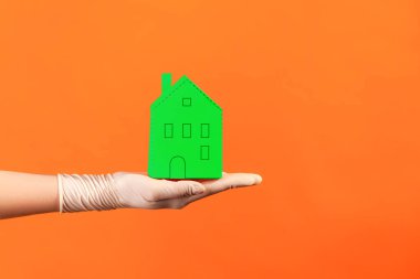 Profile side view closeup of human hand in white surgical gloves holding green paper house exterior in hand. indoor, studio shot, isolated on orange background.