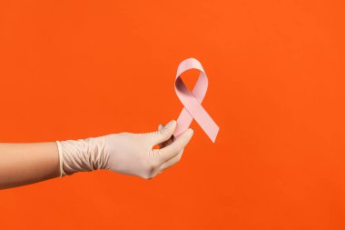 Profile side view closeup of human hand in white surgical gloves showing and holding pink ribbon, symbol of breast cancer. indoor, studio shot, isolated on orange background.