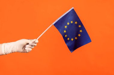 Profile side view closeup of human hand in white surgical gloves holding Eroupean union flag. indoor, studio shot, isolated on orange background.