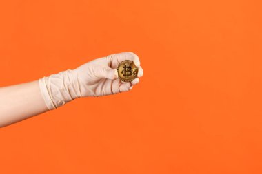 Profile side view closeup of human hand in white surgical gloves holding and showing bitcoin symbol in hand. indoor, studio shot, isolated on orange background.