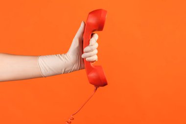Profile side view closeup of human hand in white surgical gloves holding and showing red call telephone handset receiver. indoor, studio shot, isolated on orange background.