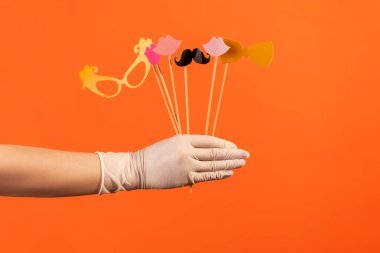 Profile side view closeup of human hand in white surgical gloves holding and showing many different stick photo-booth in hand. indoor, studio shot, isolated on orange background.