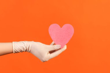 Profile side view closeup of human hand in white surgical gloves holding small pink heart shape in hand. indoor, studio shot, isolated on orange background.