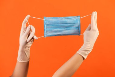 Profile side view closeup of human hand in white surgical gloves holding blue surgical medical mask. indoor, studio shot, isolated on orange background.