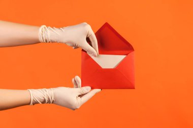 Profile side view closeup of human hand in white surgical gloves holding dargging out red opened letter envelope. indoor, studio shot, isolated on orange background.