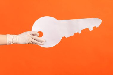Profile side view closeup of human hand in white surgical gloves holding big white key. indoor, studio shot, isolated on orange background.