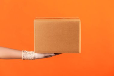 Profile side view closeup of human hand in white surgical gloves holding box. delivery concept. indoor, studio shot, isolated on orange background.