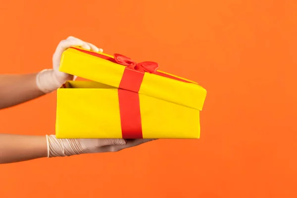 Profile side view closeup of human hand in white surgical gloves holding and opening yellow gift box. indoor, studio shot, isolated on orange background.