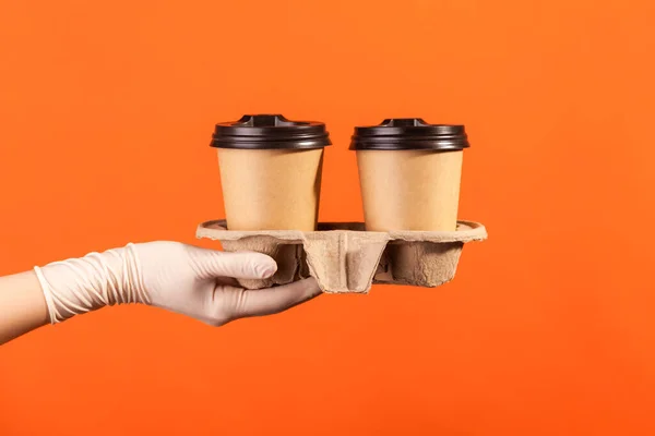 Profile side view closeup of human hand in white surgical gloves holding and showing cup of hot takeaway mug drink in hand. indoor, studio shot, isolated on orange background.