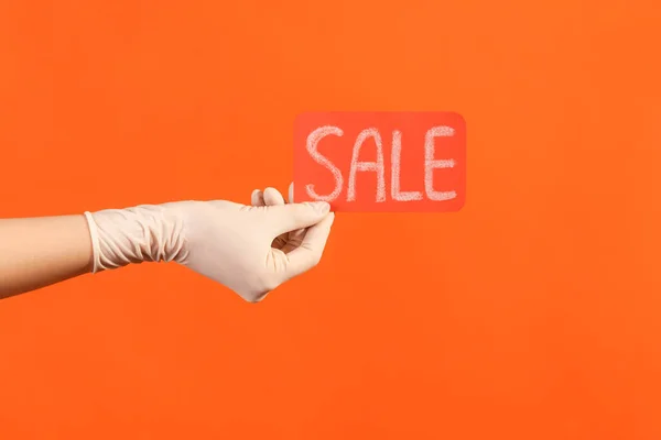 Profile side view closeup of human hand in white surgical gloves holding and showing red sale tag. indoor, studio shot, isolated on orange background.