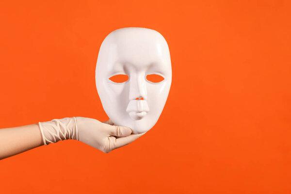Profile side view closeup of human hand in white surgical gloves holding white facial mask model. indoor, studio shot, isolated on orange background.