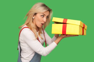 Portrait of astonished adult woman in denim overalls peeking into gift box, unpacking present, looking with interest and amazement inside, shocked by birthday surprise. indoor studio shot isolated