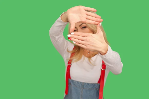 Portrait of curious adult woman in denim overalls looking through small window, photo frame shaped with fingers, focusing at target, zooming capturing idea. studio shot isolated on green background