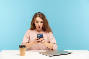 Surprised young woman, office employee, taking break at workplace with closed laptop and coffee, reading shocking message on mobile phone with amazed face, browsing web. indoor studio shot isolated
