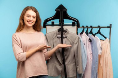Happy woman, fashion stylist standing near clothes hang on shelf in designer store, pointing at classic jacket, recommending business style, selling collection in shop boutique. indoor, studio shot