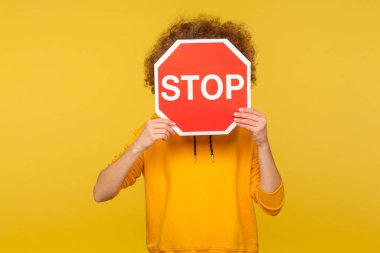 Portrait of anonymous person in urban style hoodie covering face with Stop symbol, holding red traffic sign and warning to go, denial, rejection concept. studio shot isolated on yellow background